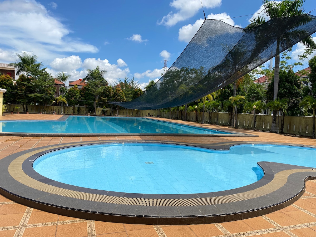 A large swimming pool area features distinct sections with a main pool and a smaller, curvy pool. Shade nets provide protection from sunlight, and landscaped greenery is visible in the background, contributing to a relaxed atmosphere.