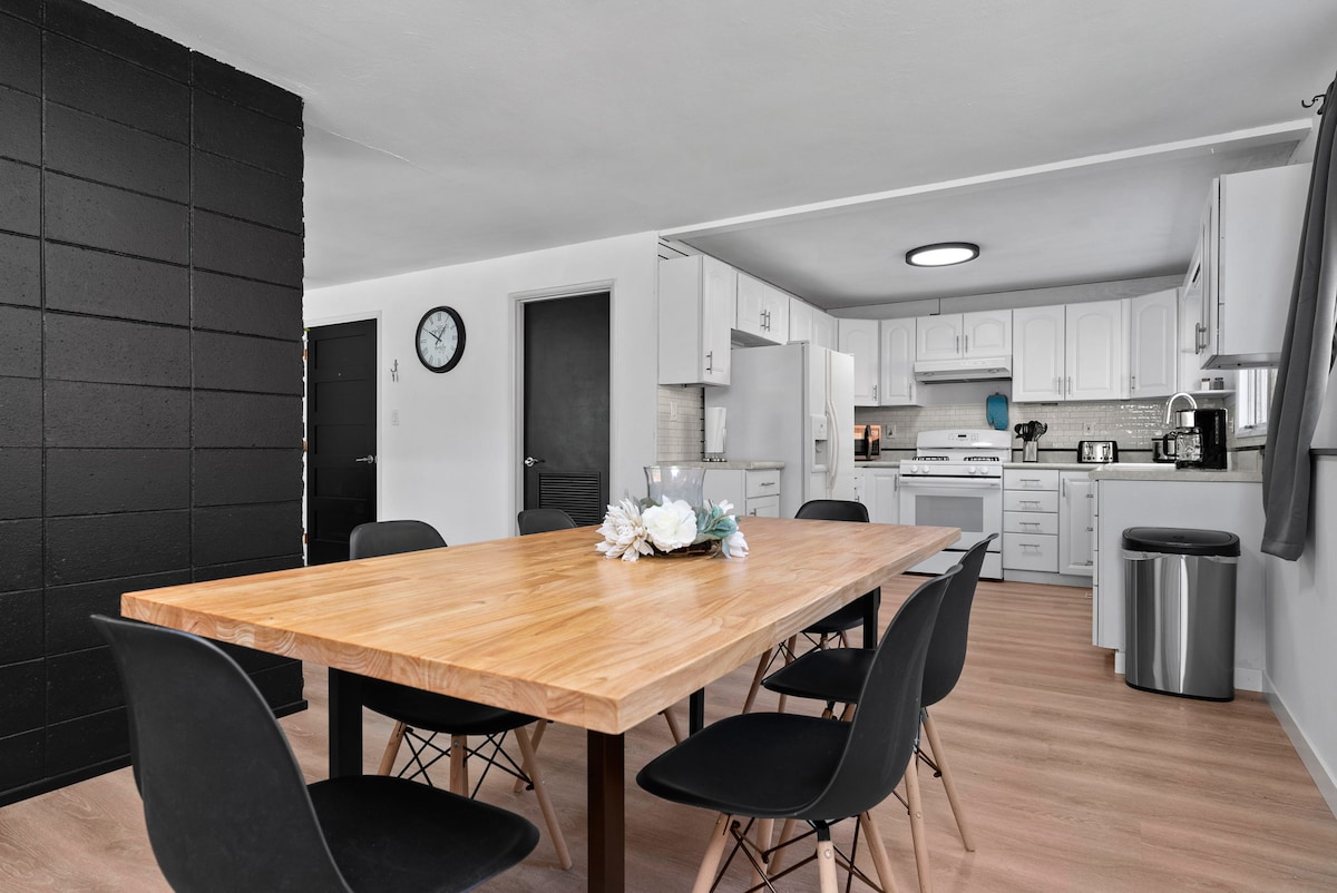 A modern dining area features a large wooden table surrounded by six black chairs. The open-concept kitchen is visible in the background, showcasing white cabinetry and stainless steel appliances. A wall clock and decorative centerpiece add subtle accents to the welcoming space.