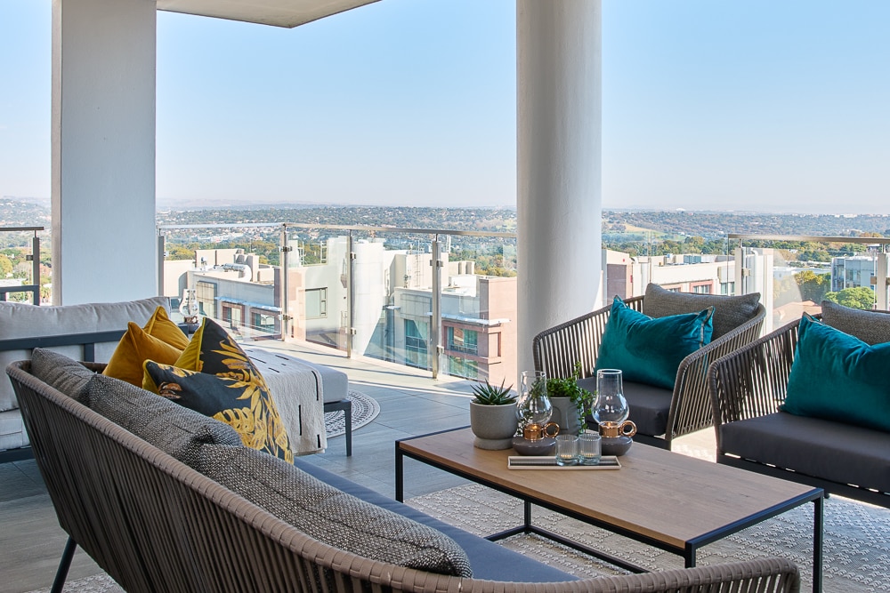 A spacious outdoor seating area is equipped with modern furniture, including a sofa and two armchairs arranged around a central coffee table. Lush cushions and a small potted plant add a touch of comfort. A panoramic view of the landscape is visible through large glass panels.