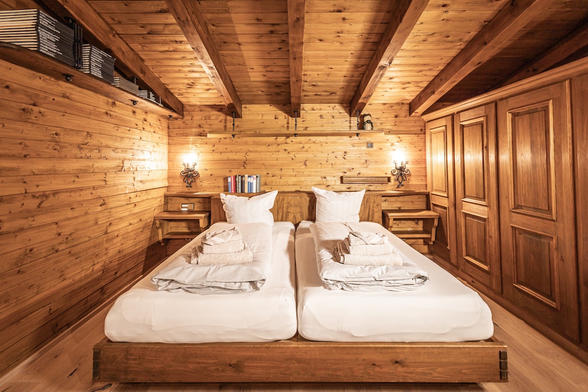 A cozy bedroom features two separate beds with light linens, positioned against warm, wooden walls. Shelves are lined with books, and bedside tables hold decorative lighting. A wooden wardrobe adds to the natural aesthetic, while exposed beams enhance the space's rustic charm.