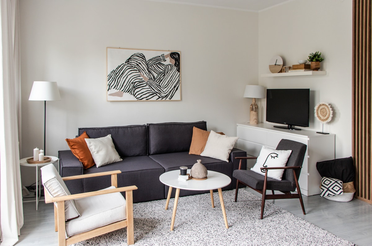 The living area features a modern, minimalist design with a dark gray sofa complemented by decorative pillows. A round coffee table sits in the center, surrounded by armchairs. Light from nearby windows highlights a wall art piece and a flat-screen television.