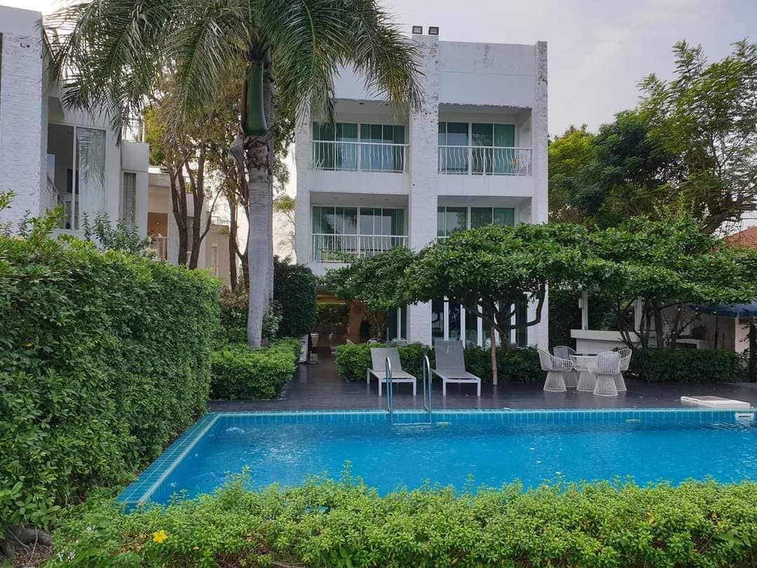 A refreshing pool is bordered by lush greenery and a well-maintained garden. Lounge chairs are positioned nearby, providing a space for relaxation. The two-story villa can be seen in the background, with balconies overlooking the tranquil outdoor area.