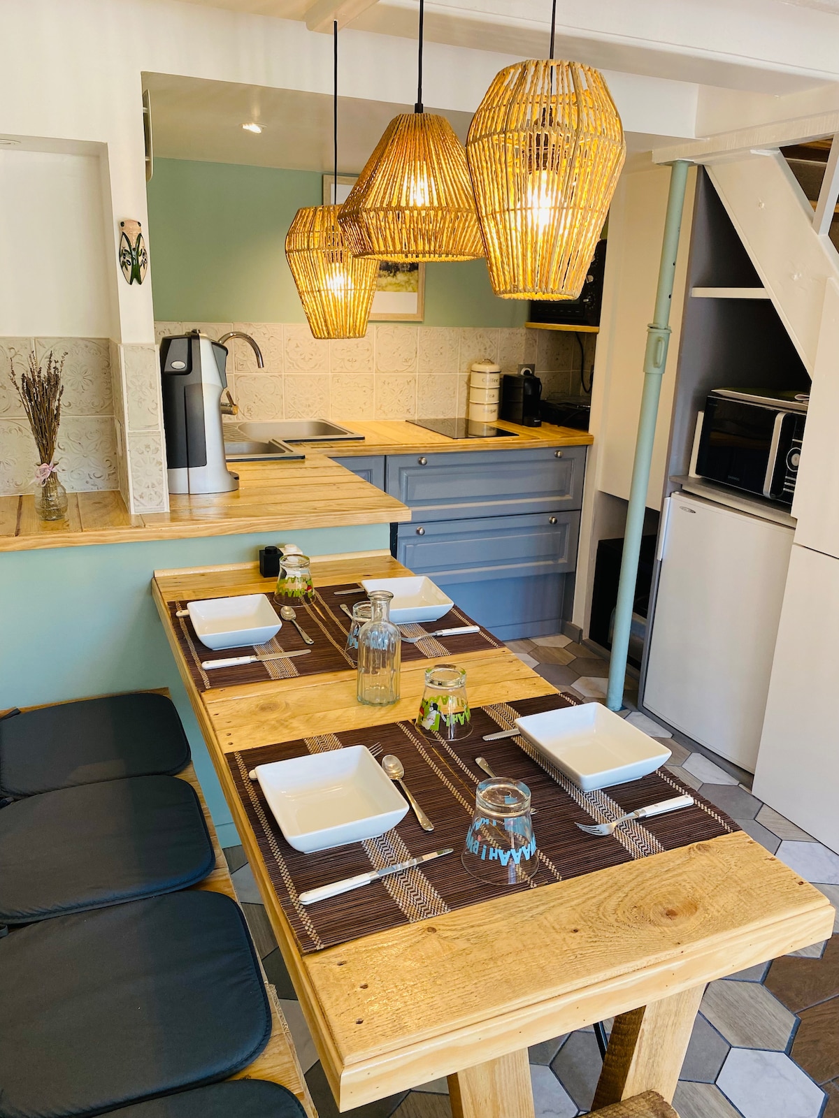 A dining area features a wooden table set for four, adorned with bowls and glassware. Three woven pendant lights hang above, illuminating the space. An open kitchen, visible in the background, is equipped with cabinetry and modern appliances.