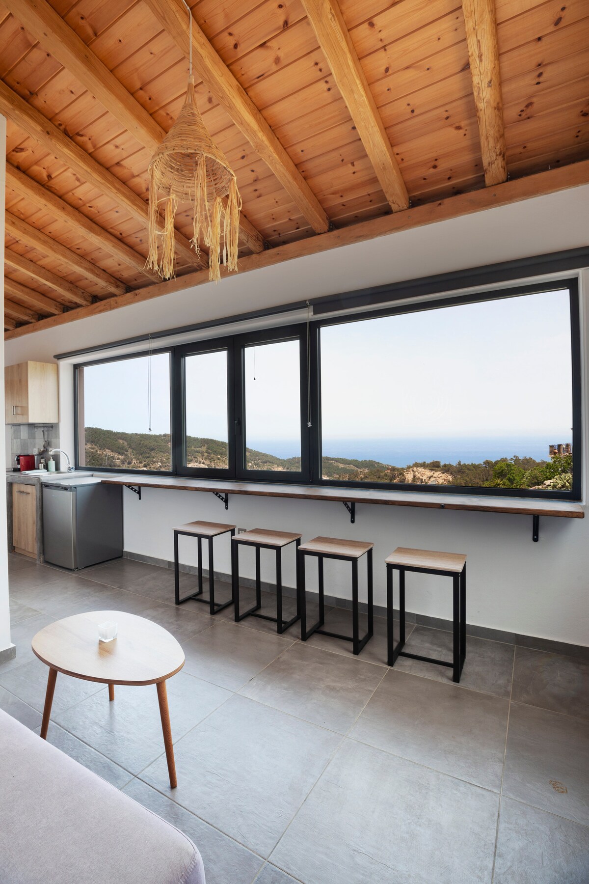 A modern kitchen area features a minimalist design, with a long counter beside large windows offering expansive views of the mountainous landscape and the sea. Four sleek bar stools are positioned along the counter, and essential kitchen appliances are visible against wooden cabinetry.