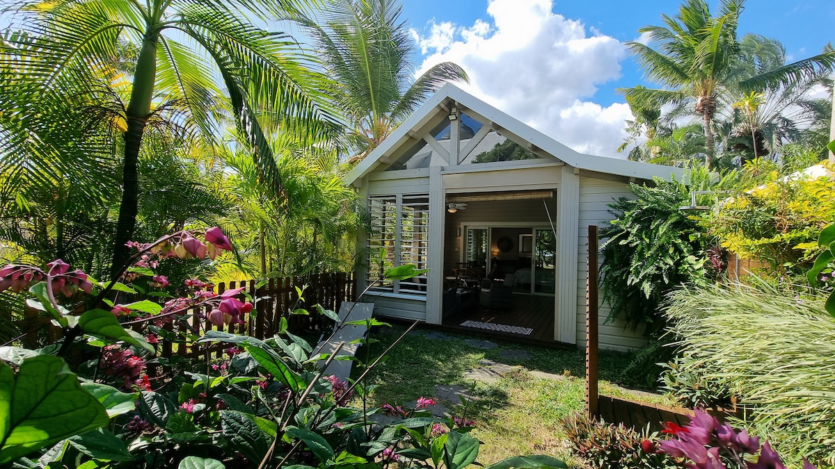 A bungalow is nestled among lush tropical foliage. Large glass windows allow natural light to fill the interior, which is visible from the garden. Plants and vibrant flowers surround the entrance, creating a serene outdoor environment that invites relaxation.