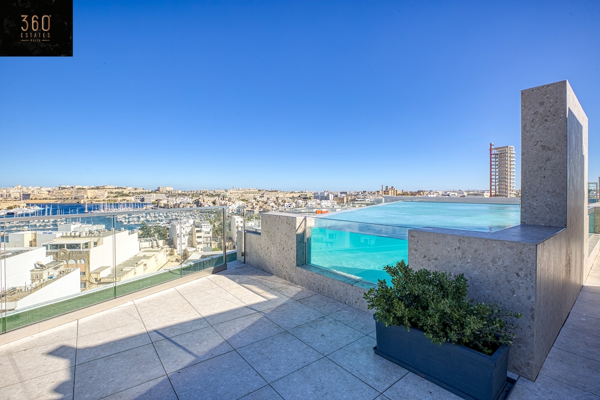 The rooftop area features a large infinity pool with stunning city and sea views. A minimalist design is highlighted by grey stone tiles and glass barriers. Potted greenery adds a touch of nature to the space, inviting relaxation in this tranquil setting.