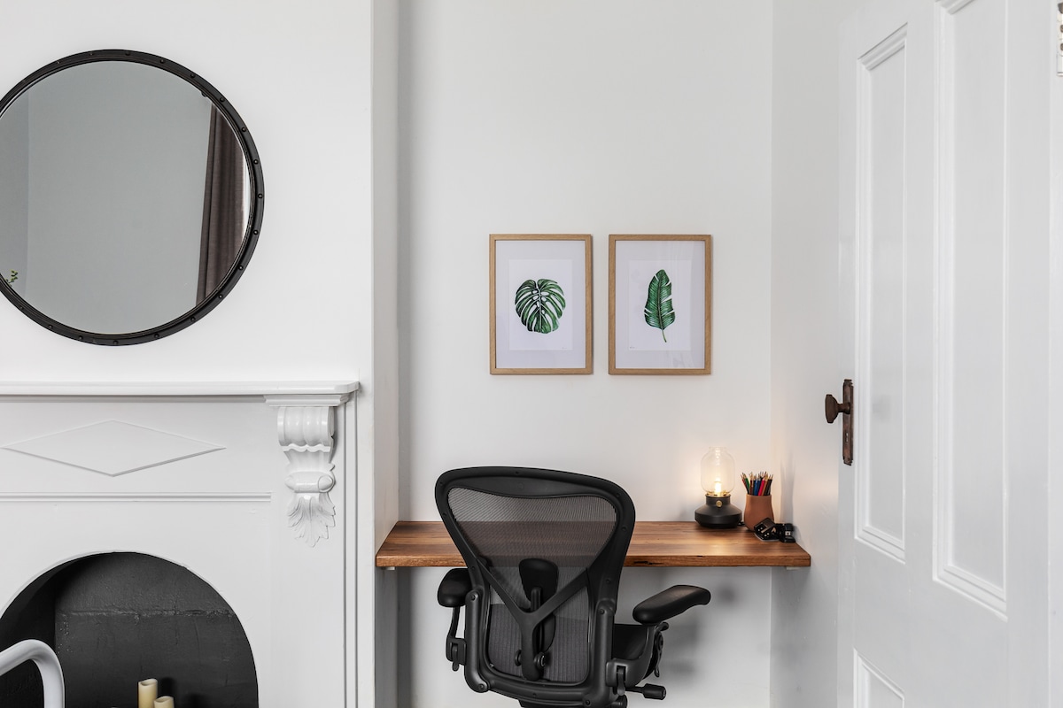 A minimalist workspace is featured in a corner, featuring a wooden desk equipped with an ergonomic chair. Two framed botanical prints adorn the wall, complemented by a circular mirror. A small candle provides soft lighting, creating a calm atmosphere for productivity.