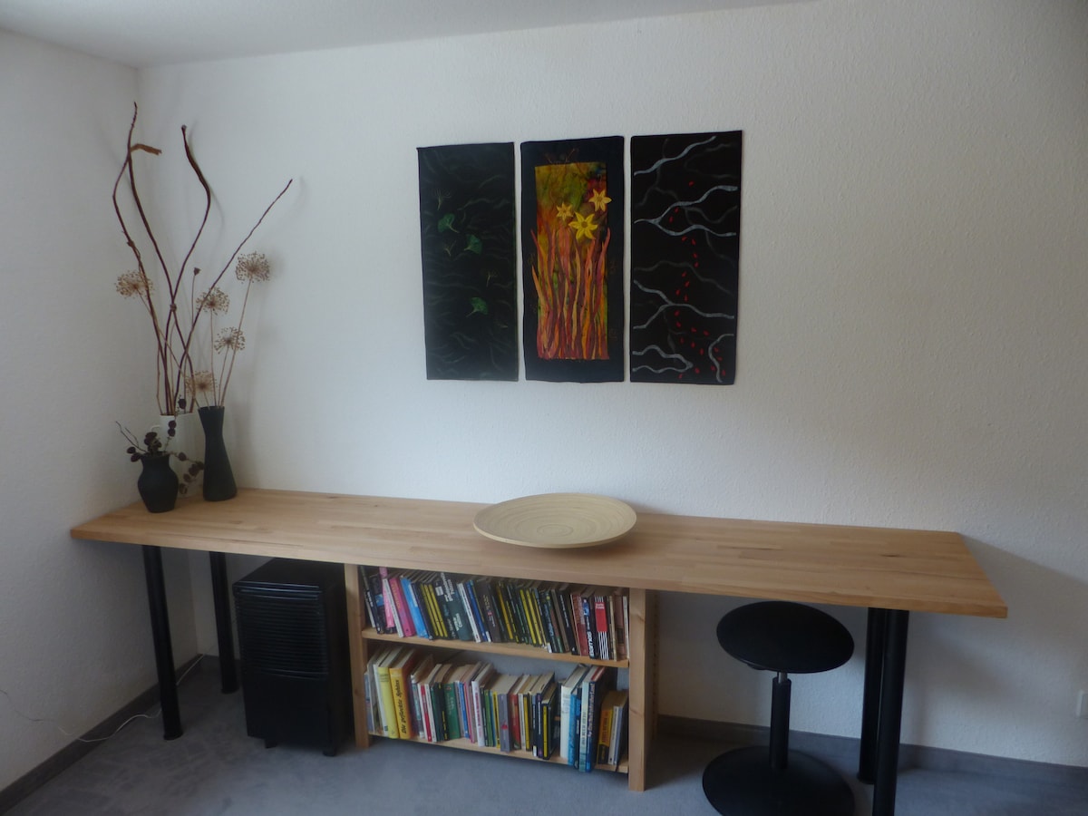 A minimalist wooden table is positioned against the wall, complemented by two black stools. Below the table, a bookshelf is filled with various books. Three framed artworks hang above, featuring vibrant colors and abstract designs.