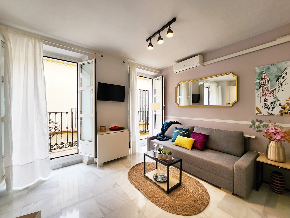 A bright living area features a gray sofa, complemented by a colorful throw pillow. A wall-mounted TV is positioned opposite. Two open balconies are seen, allowing natural light to fill the space. Decorative elements, including a low table with plants, enhance the inviting atmosphere.