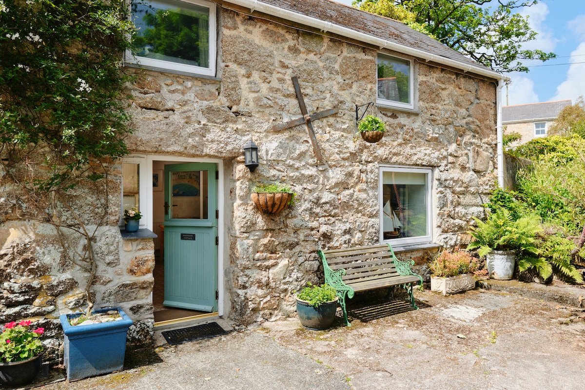 The exterior of a charming stone cottage is presented, featuring a soft green front door and several planters adorned with flowers. A wooden bench is positioned beside the entrance, while the surrounding greenery adds to the peaceful setting.