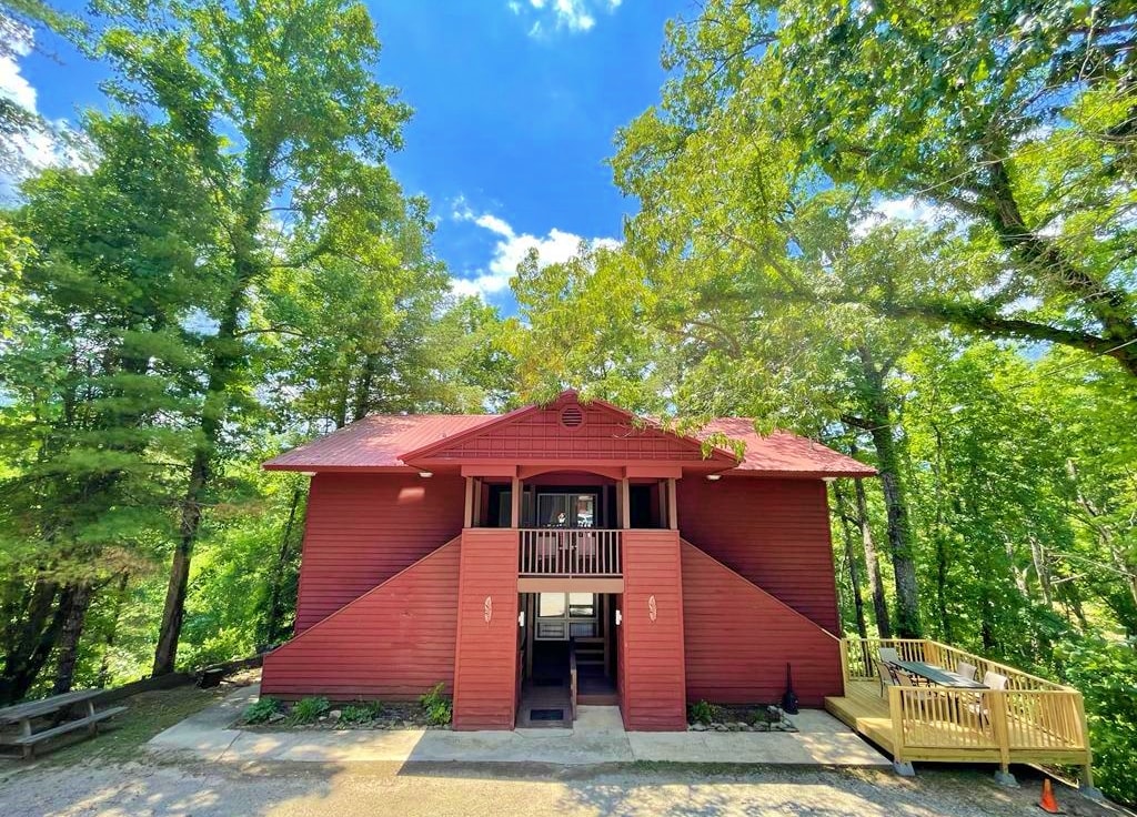 The red duplex exterior is framed by lush greenery, showcasing a two-story design with a welcoming front porch and a deck. The entrance is highlighted by a staircase leading up to the second-floor suite, set amidst a bright blue sky and gentle clouds.