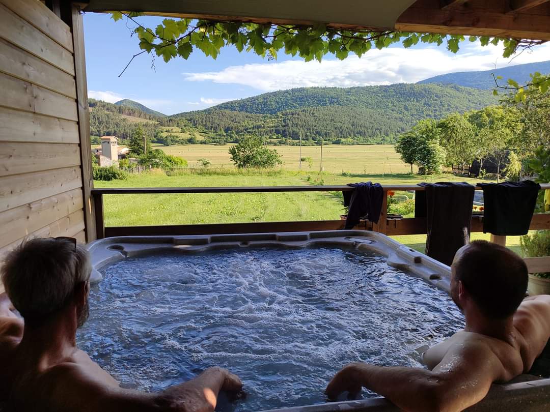 A private spa is set against a backdrop of rolling mountains and greenery. Two individuals are seated in the spa, enjoying the view from a covered space with wooden beams and climbing plants framing the scene.