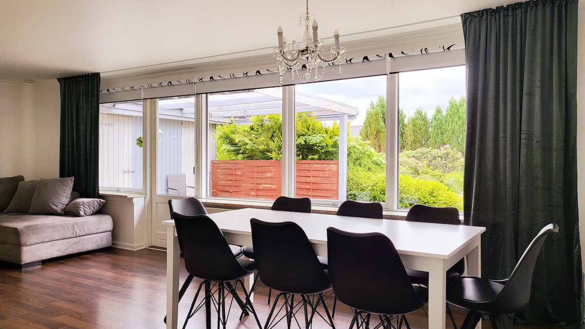 A bright dining area features a large white table surrounded by black chairs. Large windows allow natural light to fill the space, offering views of greenery outside. Elegant dark curtains frame the windows, and a chandelier hangs above the table, adding a touch of sophistication.