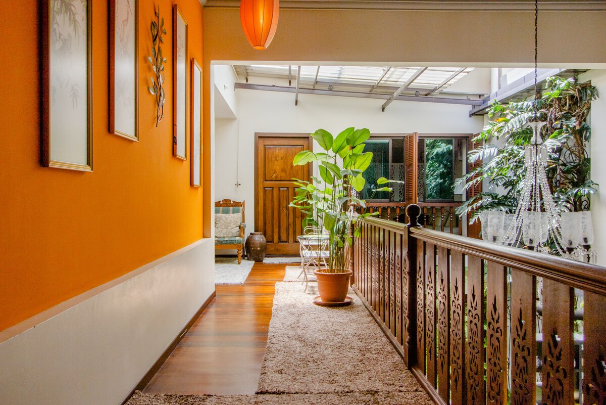 A hallway features warm orange walls adorned with framed artwork. Natural light filters through the overhead glass panels, illuminating the space, which includes a potted plant and wooden railings. The floor is covered with a plush carpet, adding to the inviting ambiance.