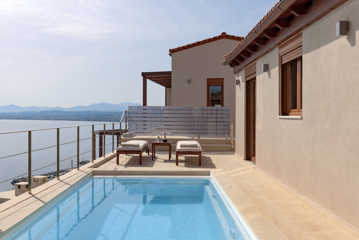 An outdoor area showcases a private pool with clear water, surrounded by two sun loungers. The villa’s exterior features neutral walls and wooden accents, with large windows allowing natural light. A balcony provides views of the sea and distant mountains.