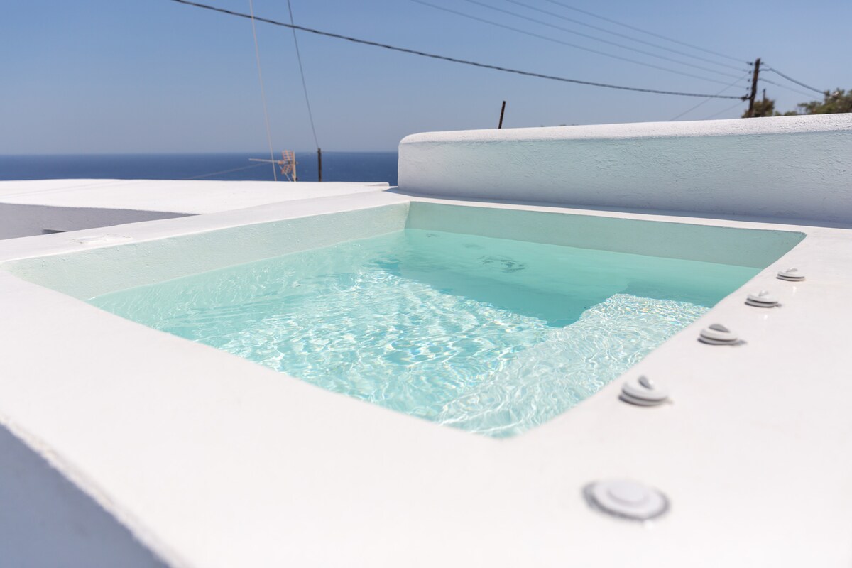 A private hot tub is set on a white patio with a smooth finish. The clear, blue water reflects the sky above, offering a serene spot for relaxation. The edge features multiple jets, enhancing the tranquil ambiance.