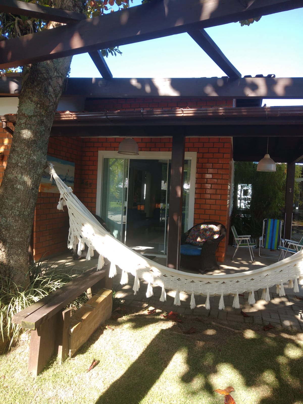 A relaxing outdoor area is highlighted by a white hammock suspended under a shaded pergola. The brick facade of the house is visible, alongside a table and chairs. Sunlight filters through the trees, enhancing the inviting atmosphere of the space.