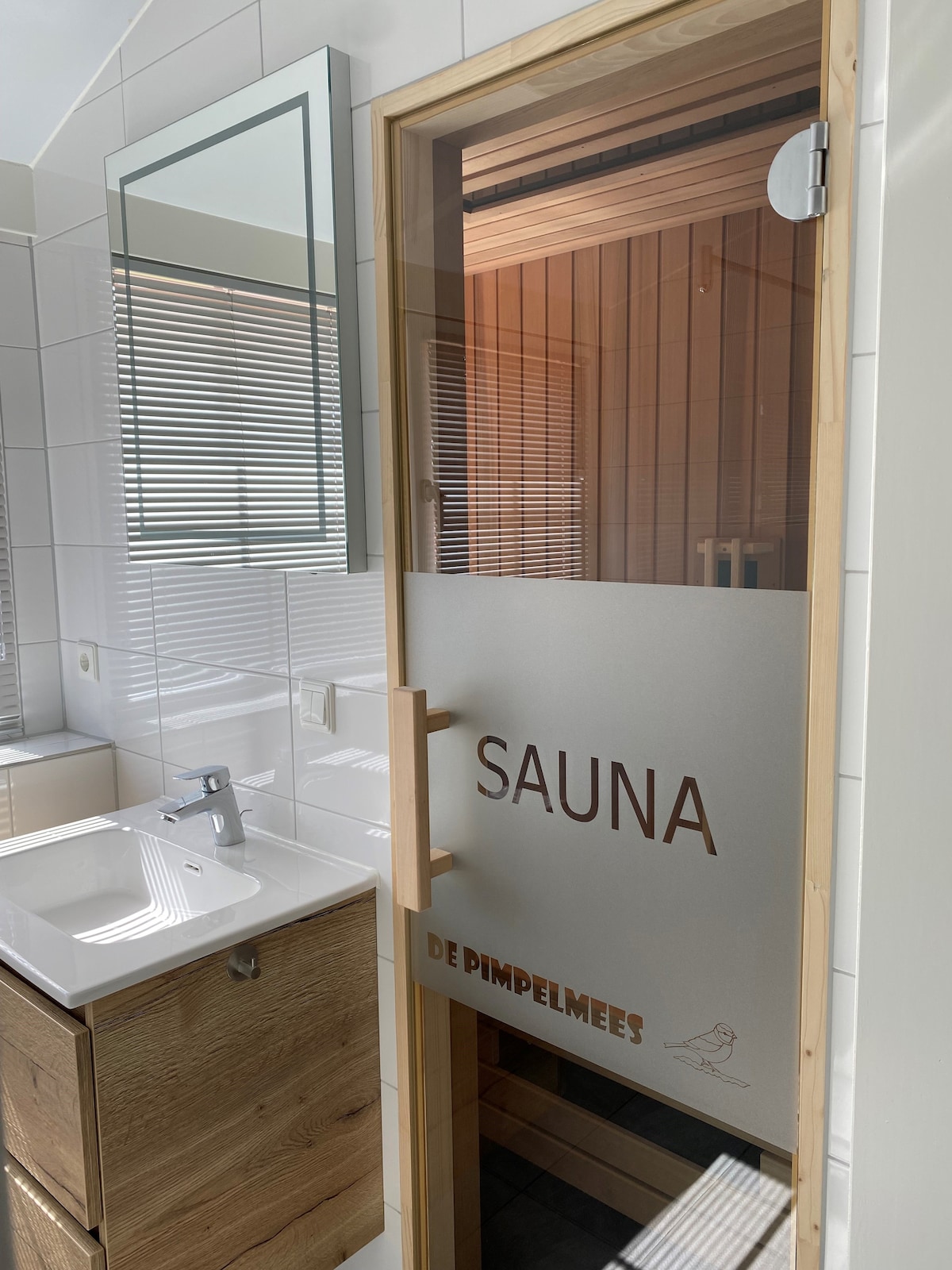 A modern bathroom is shown, featuring a sleek, minimalist sink with a large mirror above. A wooden door with frosted glass is labeled 'SAUNA', leading to a sauna area. Bright natural light illuminates the space, highlighting the clean, white tiles.