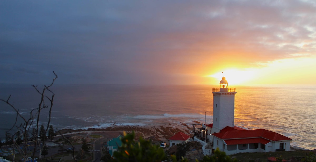 The Cape St. Blaize Lighthouse stands against a dramatic sunset, casting golden hues over the ocean. The structure features a white tower topped with a lantern, surrounded by gently rolling waves and rocky coastline. A clear sky transitions from gray to orange, enhancing the serene landscape.