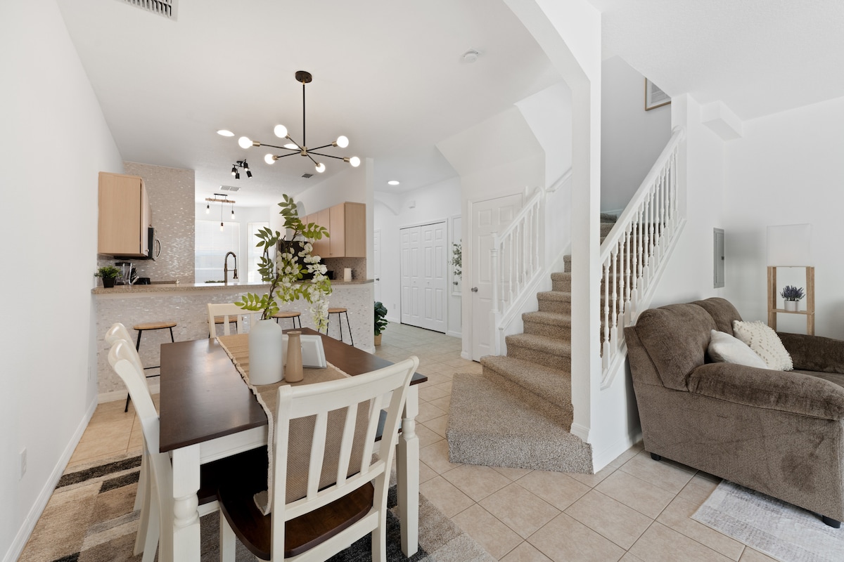An open-concept dining area features a rectangular table surrounded by four chairs, with neutral decor enhancing the space. In the background, a modern kitchen is visible, showcasing light cabinetry and stainless appliances. A staircase leads to the upper level, with a cozy couch positioned nearby.