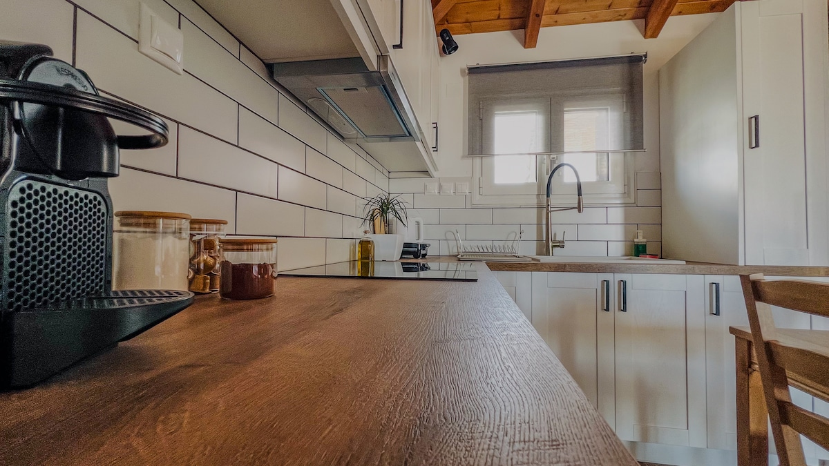 The kitchen features a wooden countertop, with a coffee machine and jars of spices. A faucet and sink are visible in the background, alongside modern cabinetry and a window that allows natural light to enter the space.