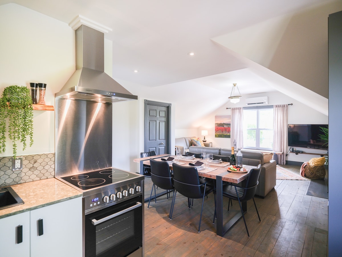A contemporary kitchen features a stainless steel stove and a dark-colored oven, paired with light countertops. An adjacent dining area is furnished with a table and chairs set for a meal. Natural light fills the space, highlighting a cozy living area with comfortable seating.