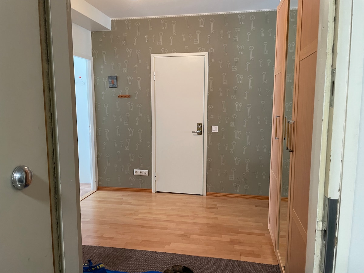 A well-lit room featuring light-colored walls with a subtle wallpaper design. The space includes a smooth wooden floor, a closed door, and a closet with sliding doors. A small wall-mounted shelf and a hook for hanging items are also visible, enhancing functional storage.