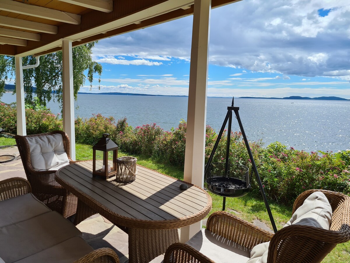 A covered outdoor seating area features a wooden table surrounded by wicker chairs. A lantern and a round grill are placed on the table, with a panoramic view of the lake visible in the background. Colorful greenery lines the edge of the water.