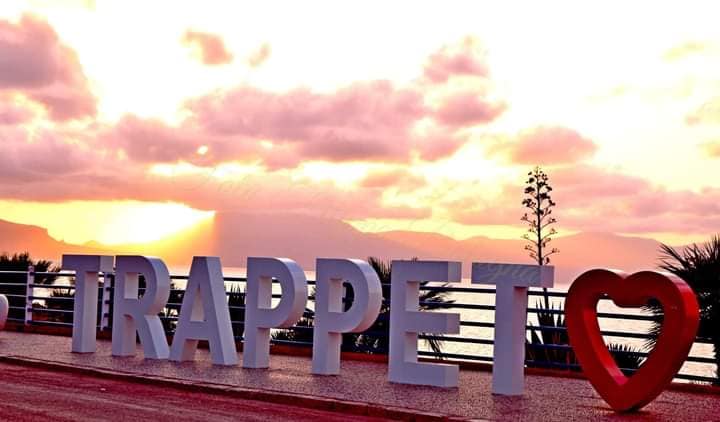A large sign displaying the word 'TRAPPET' stands prominently, complemented by a heart symbol. The scene is set against a vibrant sunset, with soft pink and orange hues filling the sky, while palm trees and distant mountains create a scenic backdrop.