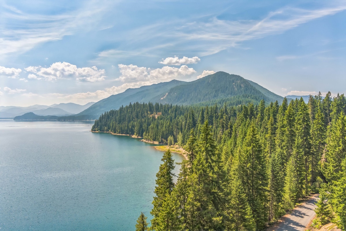 The landscape showcases a serene lake bordered by lush evergreen trees and rolling hills. The clear blue water reflects the sky, while gentle waves create a calming effect. In the distance, mountains provide a majestic backdrop under a bright, partially cloudy sky.