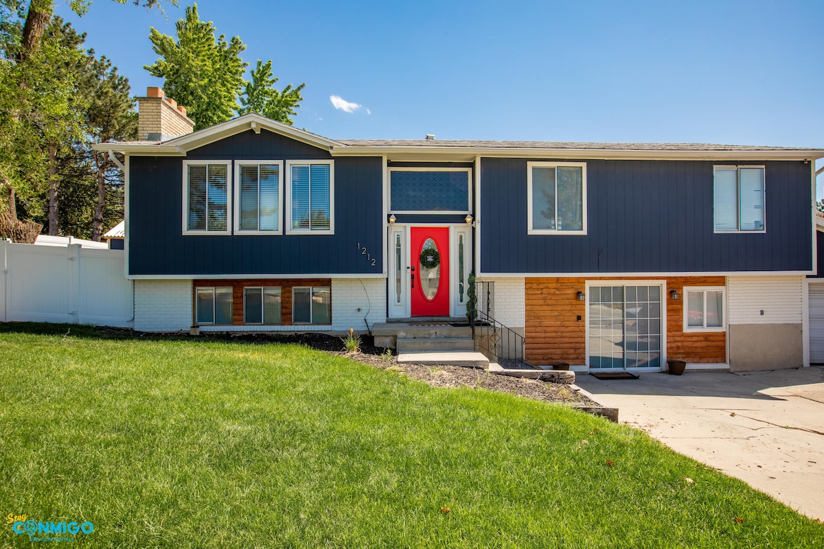 The exterior of a two-story home is displayed, showcasing a combination of blue siding and natural wood accents. A bright red front door is centered, framed by large windows on either side. Lush green grass surrounds the entrance, complemented by trees in the background.