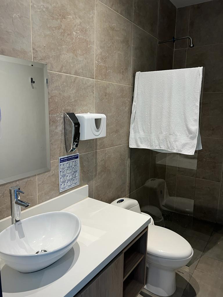 A modern bathroom is displayed, featuring a round sink atop a wooden vanity. Wall tiles in neutral tones enhance the space, while a towel hangs neatly on a rod. A shower area is visible, enclosed in clear glass, with a rainfall showerhead installed.