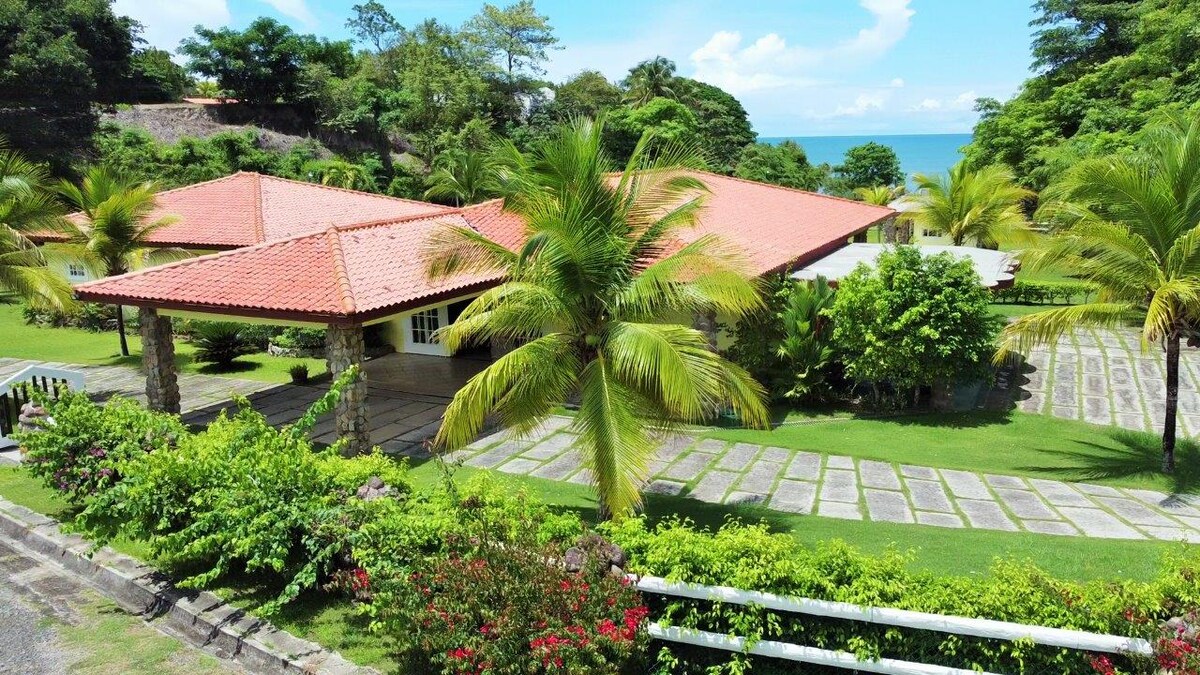The villa features a spacious layout surrounded by lush greenery, with palm trees framing the entrance. The red-tiled roof complements the vibrant tropical landscape and offers views of the nearby ocean. A pathway of stone pavers leads through the well-maintained yard.