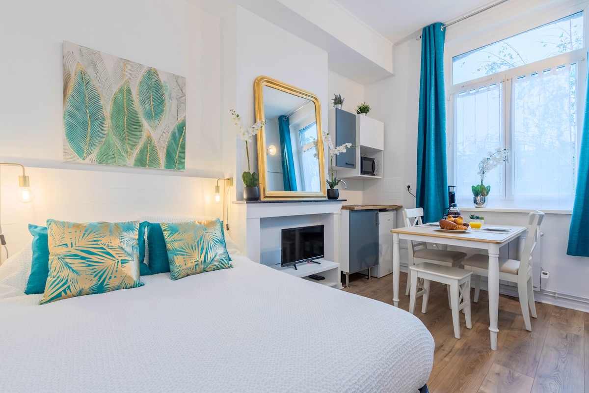 A bright sleeping area features a spacious bed adorned with decorative pillows in tropical patterns. A large golden mirror enhances the natural light reflecting from the window, while a dining table and kitchen area are located in the background.