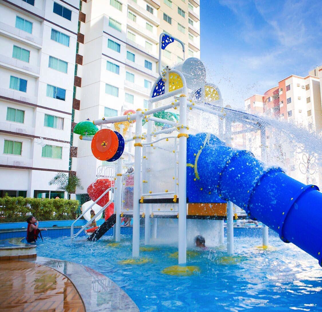A vibrant water play area features colorful slides and interactive water features within a swimming pool. Water splashes against the backdrop of a multi-story building, enhancing the fun environment. Children can be seen enjoying the various attractions under a clear blue sky.