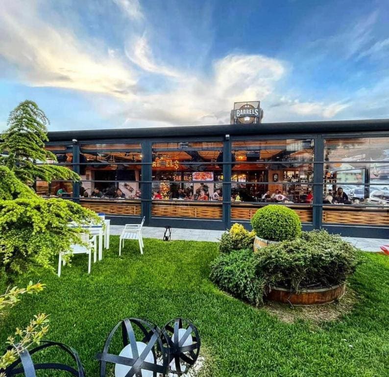 A modern outdoor dining area features a large building with expansive glass windows, allowing views of the lively interior. Lush greenery surrounds the space, with neatly trimmed bushes and white chairs arranged on the manicured lawn, inviting guests to enjoy the atmosphere.