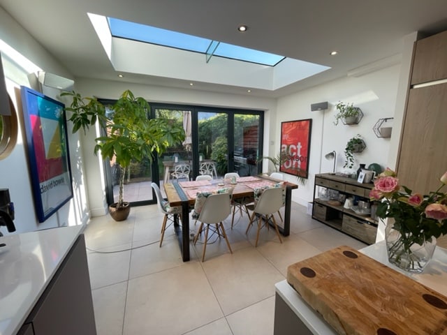 A modern kitchen/diner is illuminated by natural light coming through the skylight and bi-fold doors. A wooden dining table is surrounded by white chairs, complemented by decorative plants and framed wall art. A butcher block is visible in the foreground, and garden views are accessible through the open doors.