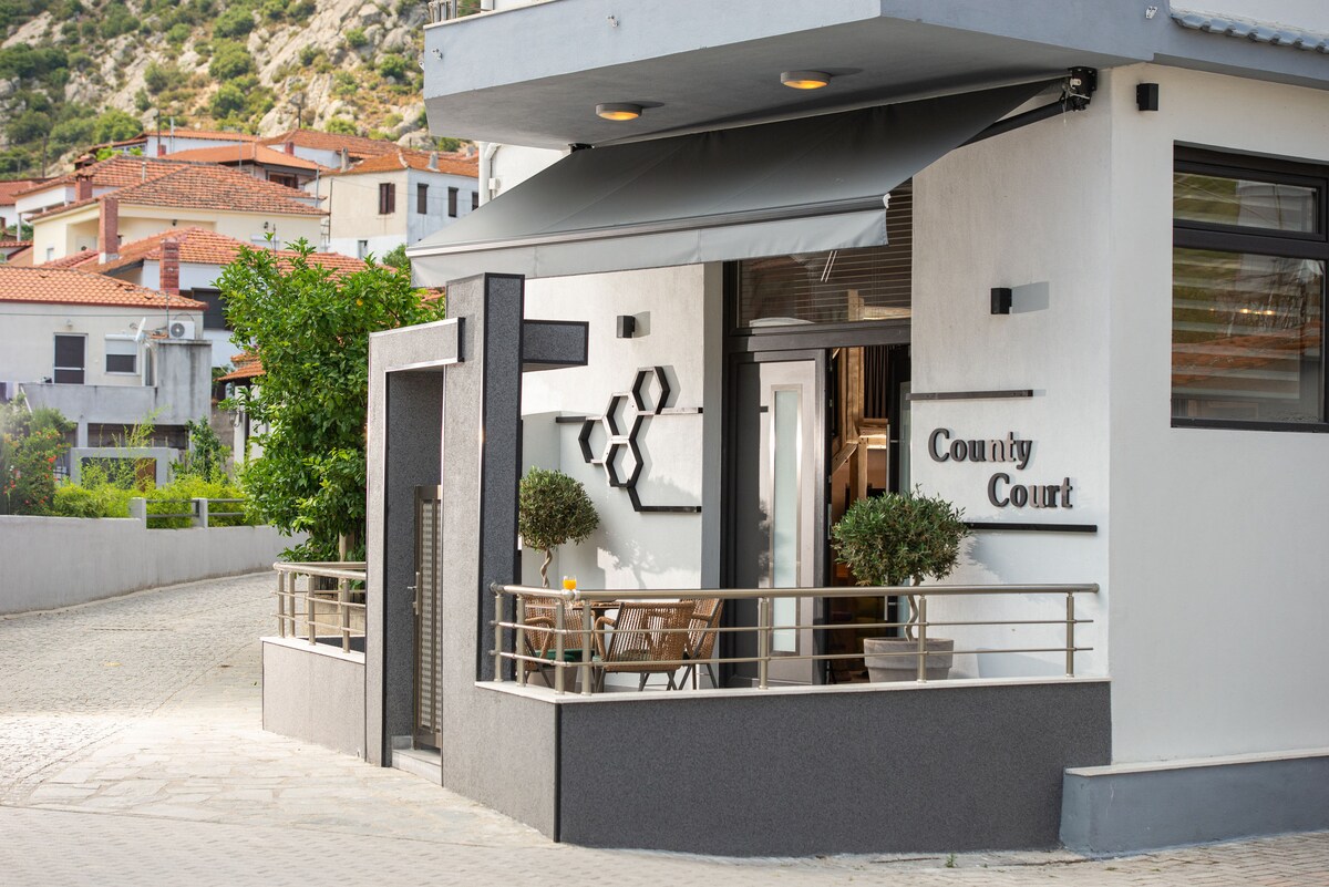 The exterior of a modern building is shown, featuring sleek gray walls and a covered terrace with seating for relaxation. Decorative plants are positioned on either side of the entrance, which is marked by the name 'County Court' in elegant lettering.