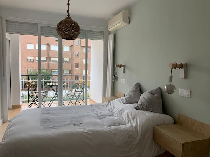 Habitación Principal. Vista luminosa a la cama de matrimonio, radiador vertical, terraza, aire acondicionado, lampara y luces empotradas. Dos mesillas de pared, una a cada lado.