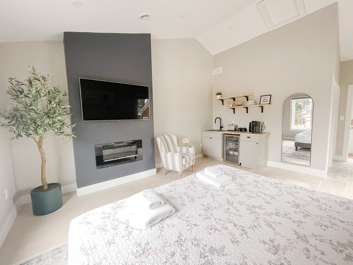 A modern loft space features a large wall-mounted television above a sleek electric fireplace. A cozy armchair sits adjacent to a kitchenette equipped with basic appliances and a mirror. Natural light filters through windows, illuminating the soft decor and comfortable bedding.