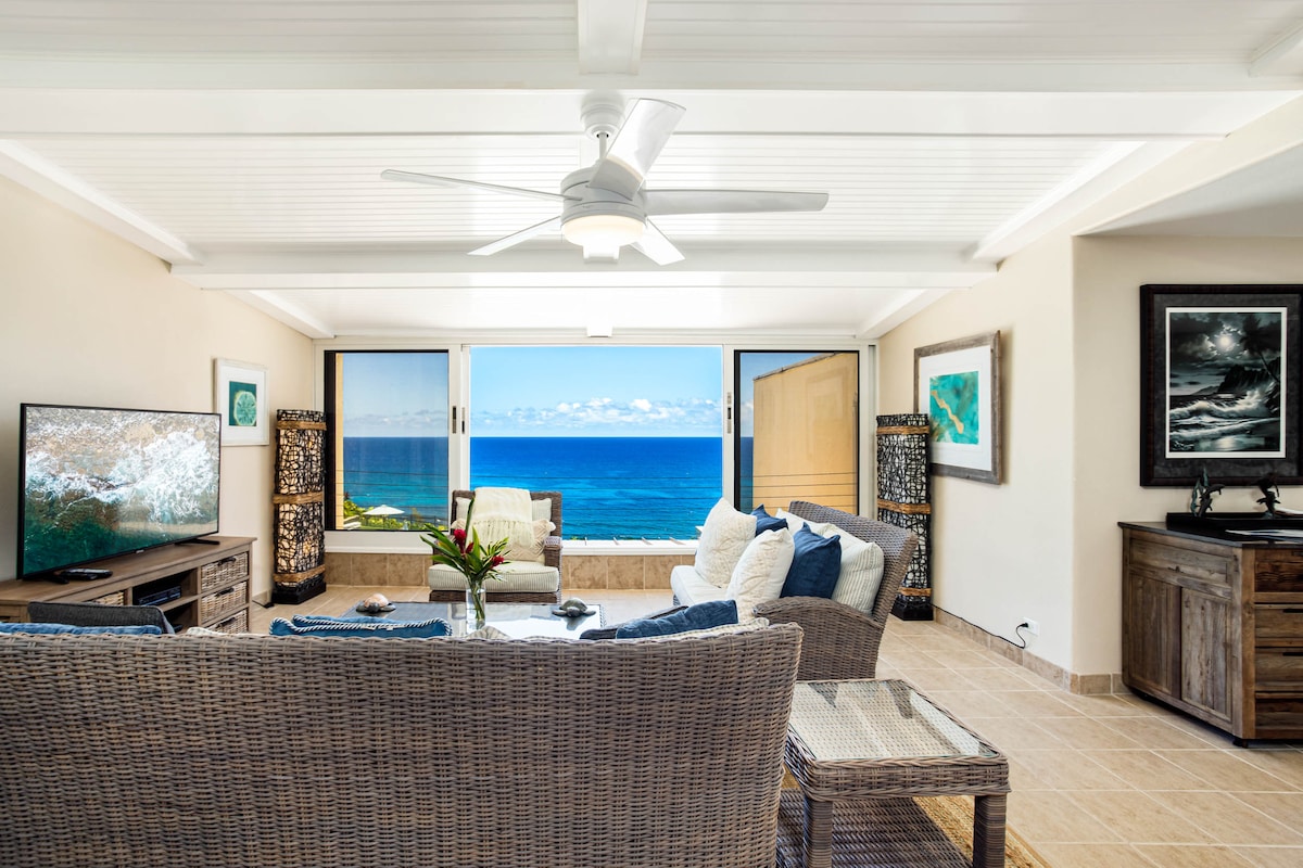 A spacious living area is furnished with modern wicker seating and a coffee table. Large windows provide unobstructed ocean views, while a ceiling fan circulates air. An HD smart TV is positioned for entertainment, and ocean-themed artwork decorates the walls.