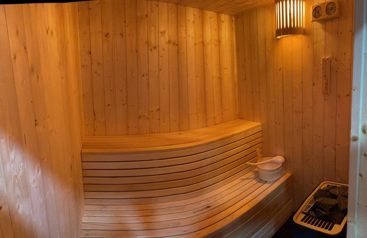 A wooden sauna room is furnished with built-in benches, creating a relaxing space. The warm wood tones are complemented by soft lighting from a wall sconce. A wooden bucket and ladle are positioned on the side, adding to the sauna's functional elements.