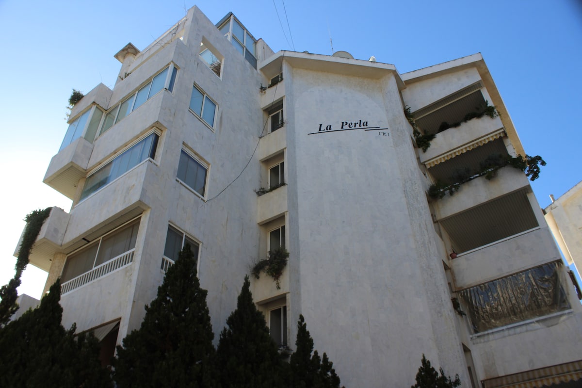 The exterior of the building, featuring a light-colored facade, displays a modern architectural design. Balconies are visible on multiple floors, adorned with greenery. A sign reading 'La Perla' is placed prominently on the upper portion, indicating the building's name.