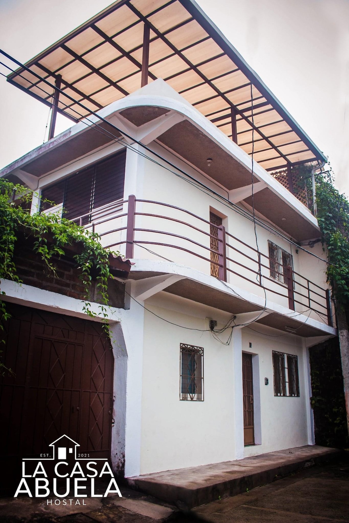 The two-story building features a clean white exterior with a covered rooftop terrace. The entrance is framed by a small staircase, while windows allow for ample natural light. Vegetation surrounds the structure, adding a touch of greenery to the setting.