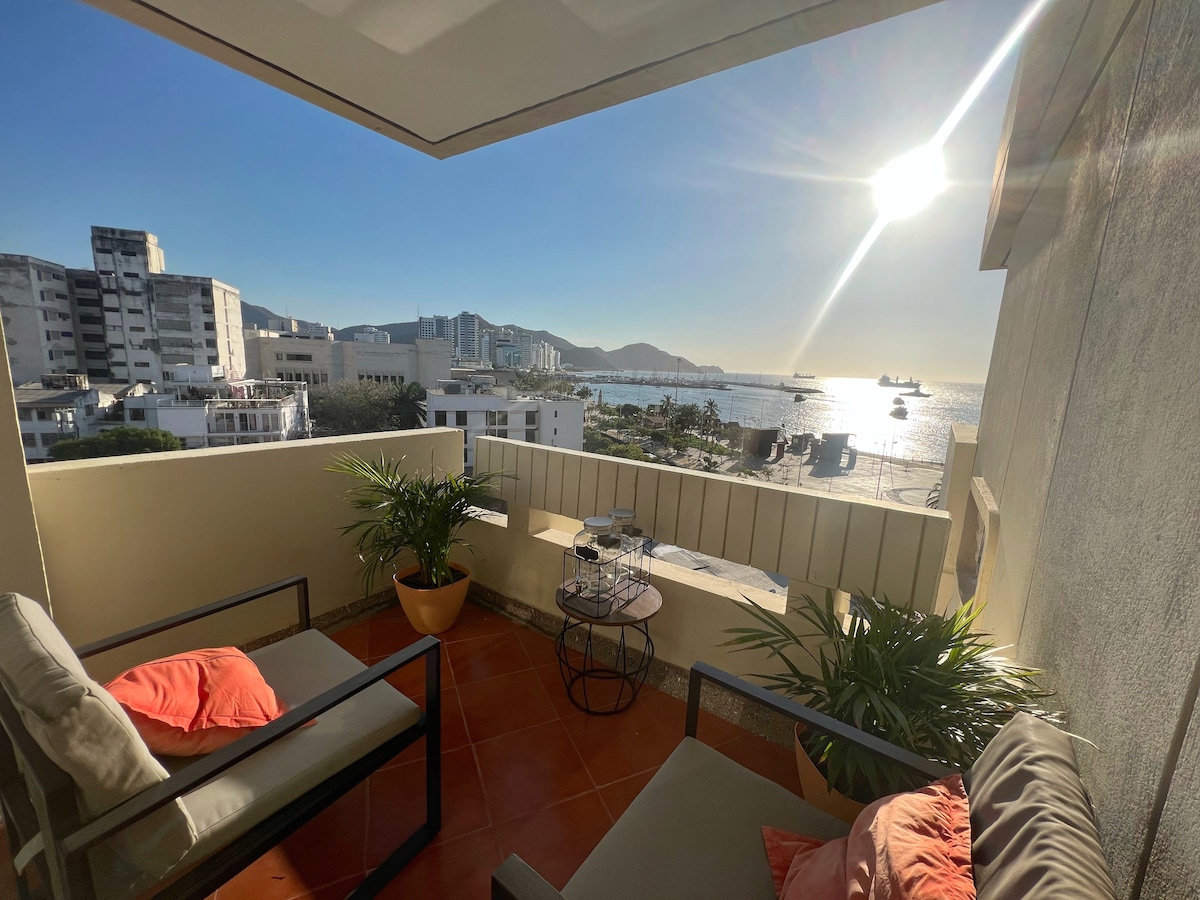 A private terrace offers a view of the bay, with sunlight reflecting off the water. Two chairs with pillows are positioned around a small table. Lush potted plants add a touch of greenery, complementing the serene outdoor space.