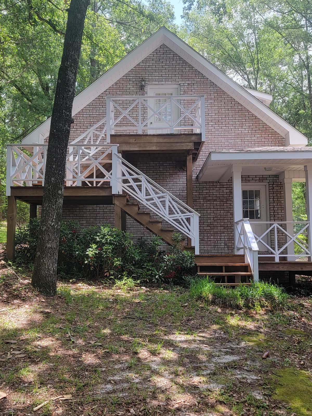 The exterior of the guesthouse is depicted, showcasing a two-story structure with brick and white railing. A set of wooden stairs leads to a covered porch, surrounded by lush greenery and trees. The natural setting emphasizes the peaceful ambiance of the location.