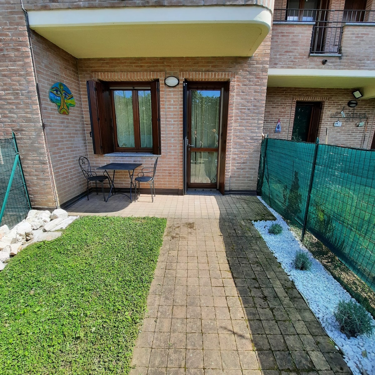 A small outdoor area features a stone-paved pathway bordered by grass and white gravel. A table and two chairs are positioned against a brick wall underneath a window with wooden shutters. A decorative wall piece adds a touch of color to the space.