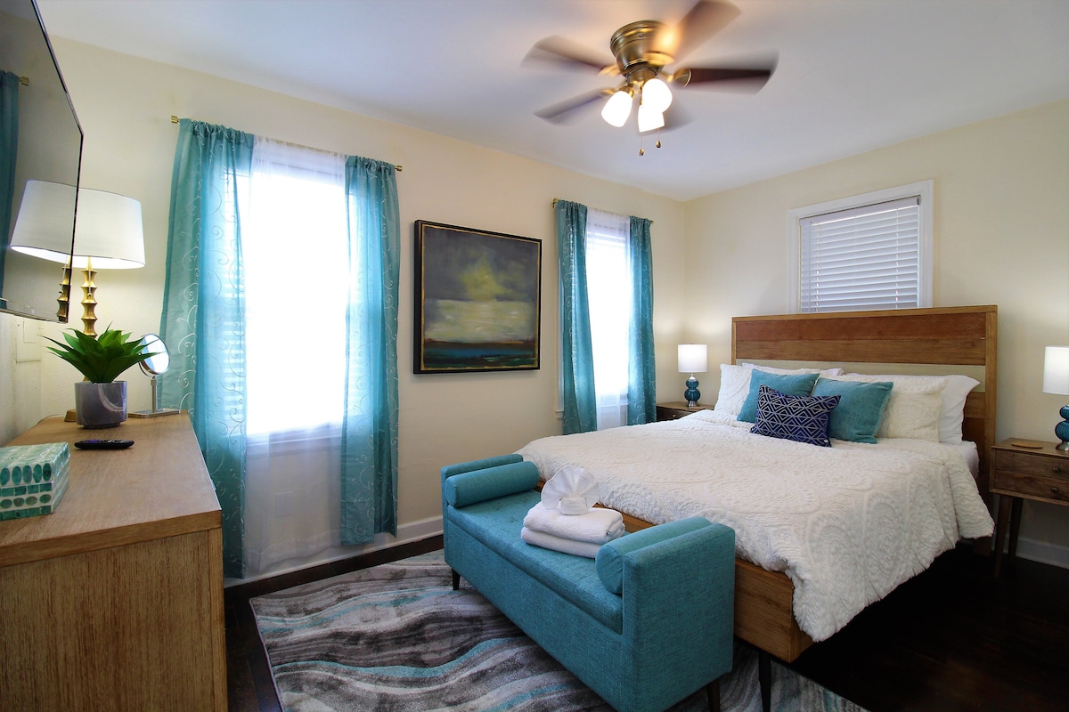 A bright bedroom features a spacious bed adorned with white and turquoise bedding. Natural light filters through two windows with sheer curtains. A dresser with a modern lamp, a painting on the wall, and a cozy seating area complete the inviting space.