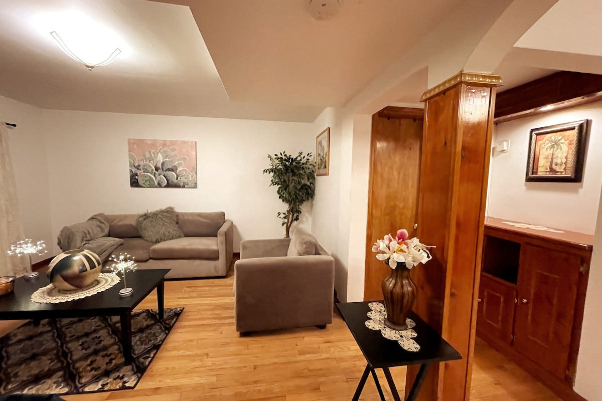 A cozy living room features neutral-toned sofas arranged around a low table. A decorative plant stands in a corner, while a piece of wall art complements the space. Natural wood accents are present in the room's surfaces, contributing to a welcoming ambiance.