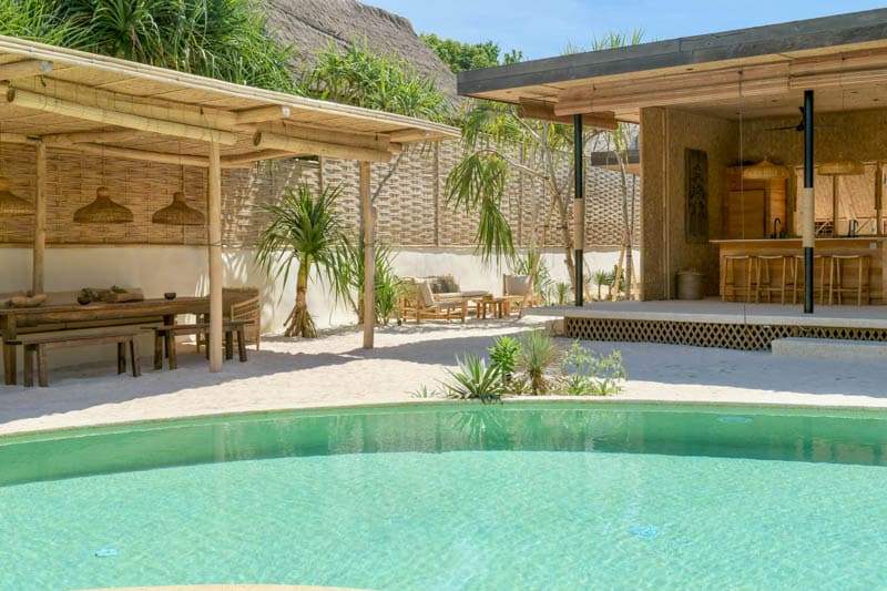 A serene outdoor space features a refreshing swimming pool surrounded by soft sand. Nearby, wooden dining tables are complemented by comfortable lounge chairs under natural shade. The open-plan living area showcases a spacious kitchen and bamboo accents, seamlessly integrating with the tropical surroundings.